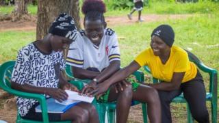 Three girls sat on green chairs outside. Hanging out and pointing at a piece of paper on one of the girls