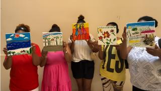 group of five girls holding up various paintings over their faces standing in front of a yellow wall