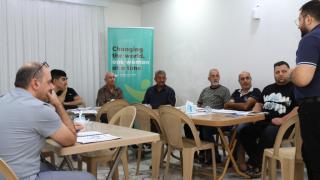 Group of men sitting around a U-shaped table all looking ahead and paying attention to a lecturer at the front of the image